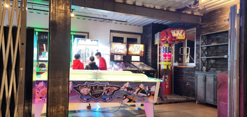 A retro arcade with various games, including pinball and a claw machine, and people enjoying the space.